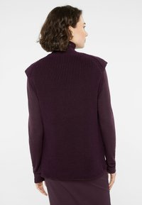 Ärmellose lila Strickweste mit hohem Kragen, strukturierter Rückseite und glattem Langarmoberteil darunter. Schlichtes Design, figurbetonte Silhouette.