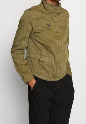 Person trägt eine olivgrüne, knöpfbare Jacke mit Klappentaschen und schwarze Hose, Hände in den Taschen, leicht nach links schauend.