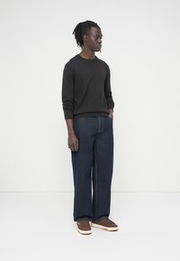 Άνδρας στέκεται σε λευκό φόντο φορώντας μαύρο μπλουζάκι με μακριά μανίκια, άνετα σκουρόχρωμα τζιν και καφέ αθλητικά παπούτσια με λευκές σόλες.