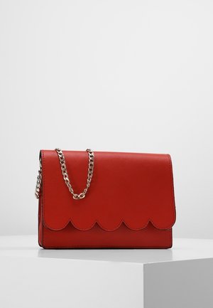 Borsa a tracolla in pelle rossa con patta a bordo smerlato e tracolla a catena color argento, posata su una superficie bianca con sfondo chiaro.