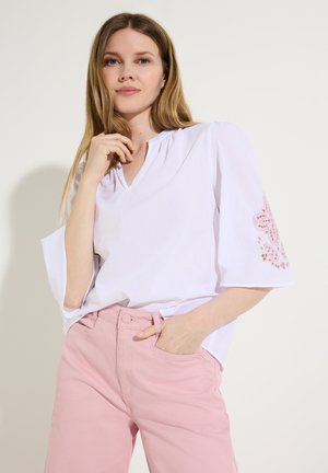 Frau mit langen Haaren, die eine weiße Bluse mit pinker Stickerei an den Ärmeln und hellrosa Hose trägt, posiert mit einer Hand in der Tasche.