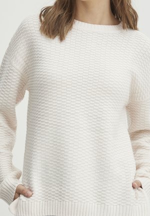 Weißer strukturierter Pullover mit lockerer Passform, ausgestattet mit einem quadratischen Strickmuster, gerippten Bündchen und einem runden Halsausschnitt. Seitentaschen sind sichtbar.