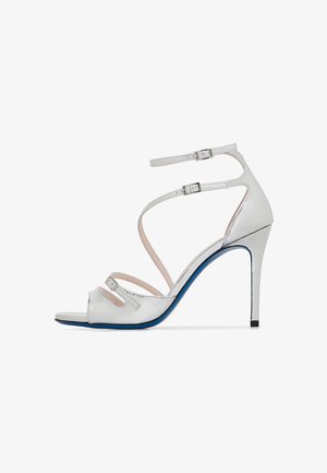 White leather high-heeled sandal with crisscross straps, an open toe, a blue sole, and two adjustable ankle buckles.