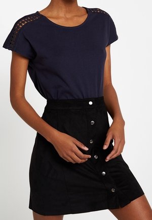 Vrouw draagt een marineblauw kortemouwen top met kanten details op de schouders en een zwarte hoge taille rok met knopen aan de voorkant.