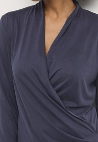 Kvinna som bär en långärmad, marinblå omgjord topp med V-ringning, som visar övre bröstet och axlarna.