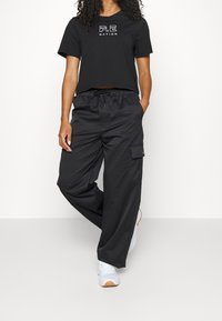 Black cropped T-shirt with white "P.E. NATION" logo, paired with loose black cargo pants featuring side pocket and drawstring waist. White sneakers.