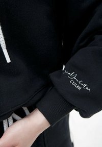 Czarna bluza z kapturem i ściągaczem, z prążkowanymi mankietami. Posiada białe, kursywne napisy "seeYouLater" oraz "CUL8R" na rękawie. Miękka tekstura materiału.