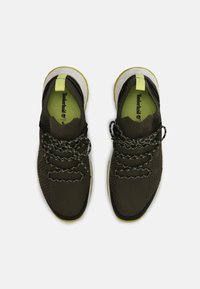 Baskets tricotées vert foncé avec une tige texturée, des lacets réfléchissants, des accents jaune vif et une semelle en caoutchouc blanche.