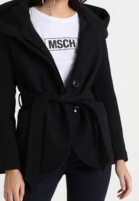 Mujer con un abrigo negro ceñido en la cintura sobre una camisa blanca con el texto "MSCH", combinada con pantalones oscuros, de pie contra un fondo claro y liso.