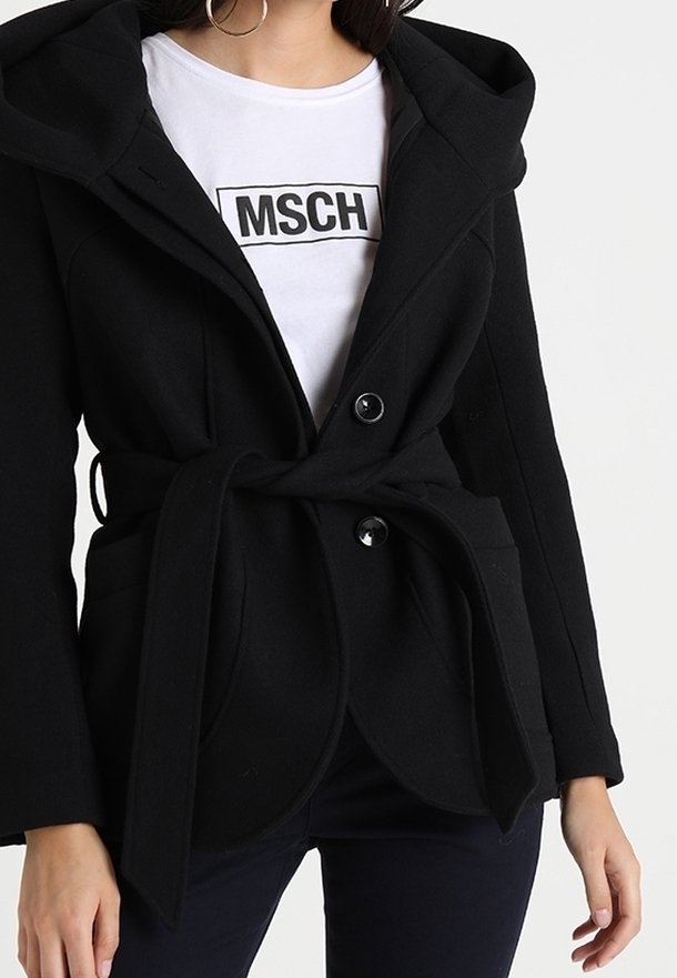 Mujer con un abrigo negro ceñido en la cintura sobre una camisa blanca con el texto "MSCH", combinada con pantalones oscuros, de pie contra un fondo claro y liso.