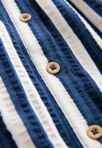 Navyblaue und weiße gestreifte Stoff mit Texturvariationen. Die Knöpfe sind rund, hellbraun und haben ein Lochdesign mit vier Löchern.
