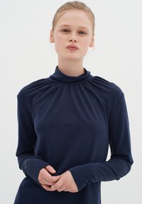 Námornícky dlhý rukáv s vysokým golierom, s naberanými detailmi na krku a priliehavými manžetami, vyrobený z hladkej, elastickej látky.