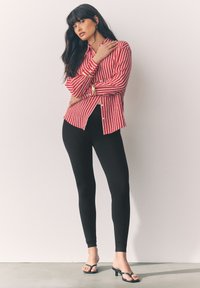 Rød og hvidstribet skjorte med knapper og lange ærmer, kombineret med sorte leggings og sorte hælede sandaler, sat mod en neutral baggrund.