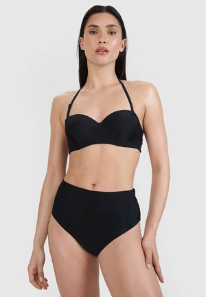 Kobieta z długimi ciemnymi włosami, ubrana w czarne bikini z wysokim stanem i górę bikini bez ramiączek z paskiem halter, na szarym tle.