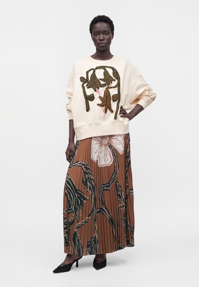 Femme debout, main sur la hanche, portant un sweat crème avec broderie florale marron et une jupe plissée marron avec un grand imprimé floral et des talons noirs.