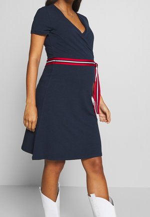 Femme enceinte portant une robe bleu marine jusqu'aux genoux avec une ceinture rayée rouge et blanche, assortie à des bottes blanches montantes jusqu'aux genoux.
