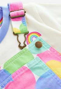 Värikrika hängselbyxor med pastellfärgade prickar, blå krage, metallbeslag och regnbågsbroderi, som visar en livfull textur och design.