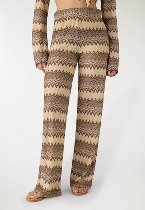 Donna che indossa pantaloni larghi beige e marroni a motivo zigzag con top a maniche lunghe abbinato e sandali color cuoio.