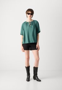 Teal t-paita, jossa on avainaukko, yhdistettynä mustiin shortseihin ja mustiin cowboy-bootseihin, joissa on monimutkaista ompelua.