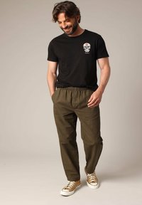 T-shirt noir avec un graphisme de skull, associé à un pantalon vert olive à taille élastique et des baskets beiges avec des semelles blanches.