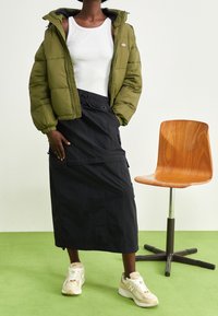 Giacca a vento verde oliva, canotta bianca, gonna midi nera con tasche laterali e sneakers beige con logo New Balance. Sedia di legno sullo sfondo.