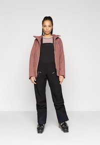 Ūdensizturīga rozā jaka ar kapuci, kombinēta ar melnajām kombinezoniem un melniem slēpošanas zābakiem. Jaka ir ar rāvējslēdzēju un teksturētu paneļu akcentiem.