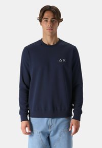 Giovane uomo che indossa una felpa blu navy con un logo a triangolo e X sul petto e jeans azzurri, in piedi di fronte a uno sfondo semplice.