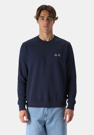 Giovane uomo che indossa una felpa blu navy con un logo a triangolo e X sul petto e jeans azzurri, in piedi di fronte a uno sfondo semplice.