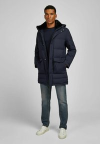 Marineblauwe pufferjas met een afneembare capuchon, voorrits, twee klepzakken en een zachte binnenvoering, gecombineerd met denim jeans en witte sneakers.