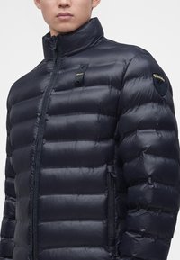 Homme portant une doudoune matelassée noire avec col montant, fermeture éclair devant, poche latérale zippée et écussons de marque sur la poitrine et la manche.