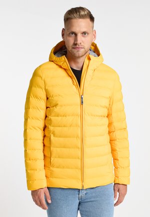 Gele gewatteerde jas met een capuchon, voorzien van gestikte secties, een voorkant met rits en een grijze binnenvoering, gedragen over een zwart shirt.