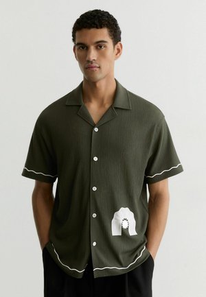 Mees kannab lühikeste varrukatega oliivrohelist särke, millel on valged lainelised äärised ja alumisel esiküljel valge graafika, taust on ühevärviline.