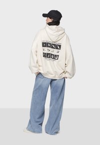 Sudadera crema con capucha, con un estampado gráfico de ojos y texto en la parte de atrás. Combinada con unos vaqueros de pierna ancha en azul claro. Lleva una gorra negra.