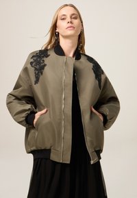 Giacca bomber verde oliva con ricami floreali neri sulle spalle, chiusura con zip davanti, polsini elastici e tasche laterali. Tessuto liscio e lucido.