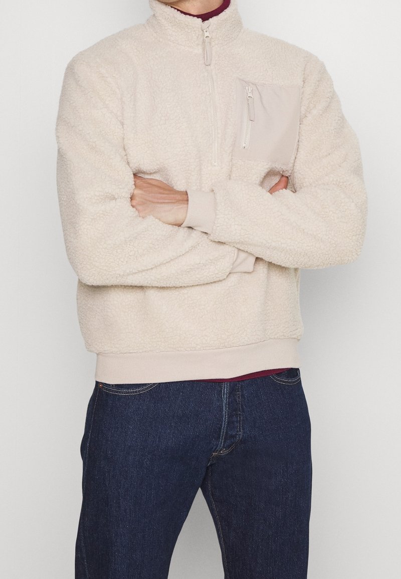 Persona vestida con un pulóver de forro polar beige con un bolsillo en el pecho con cremallera y jeans azul oscuro, de pie con los brazos cruzados.