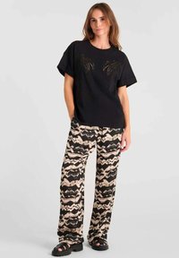 Camiseta negra de manga corta con bordado de palmera, combinada con pantalones beige holgados con un patrón ondulado negro y sandalias negras gruesas.