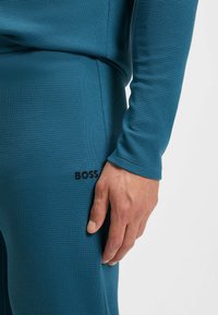 Gros plan sur la main d'une personne reposant près d'un pantalon texturé bleu avec le logo noir BOSS brodé près de la poche.