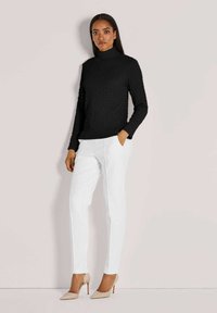 Frau, die an einer Wand steht, gekleidet in einen schwarzen strukturierten Rollkragenpullover, weißen maßgeschneiderten Hosen und beigen High-Heels, mit einer Hand in der Tasche.