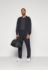 Tenue sportive en tissu bleu marine foncé, comprenant une veste zippée, un pantalon assorti et un sac de sport noir. Des baskets blanches complètent le look.