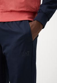 Veste de sport rouge et marine en tissu texturé. Le pantalon marine a une finition lisse et présente le logo EA7 sur la jambe gauche. Comprend un détail de poche.