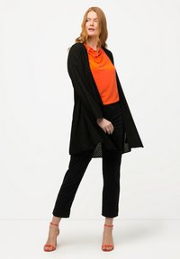 Svart texturerad öppen cardigan över en orange koftahals topp, kombinerad med svarta cropped byxor och röda högklackade sandaler.