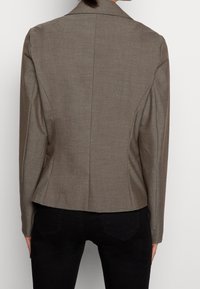 Ruskea mittatilauspuku, jossa on jämäkkä muotoilu ja notched-lapels. Sisältää hienovaraisen tekstuurin, viistoja saumoja ja istuvan siluetin.