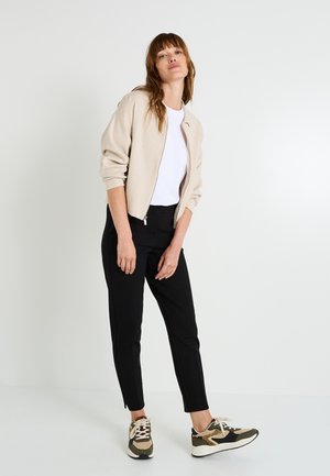 Pull beige à fermeture éclair, t-shirt blanc, pantalon noir avec fente latérale, et baskets vertes/marron avec accents à motifs. Textures lisses partout.