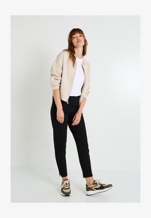 Jersey beige con cremallera, camiseta blanca, pantalones negros con abertura lateral y zapatillas verdes/marrones con detalles estampados. Texturas suaves en todo el conjunto.
