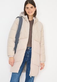 Ung kvinde iført en beige quilted jakke med hætte, blå jeans og en grå rygsækstrap over skulderen, smilende til kameraet.