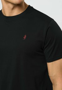 Vīrietis, kurš valkā melnu t-kreklu ar apaļu kaklu un mazu sarkanu izšūtu logotipu uz kreisā krūtīm, gaiši pelēkā fona.