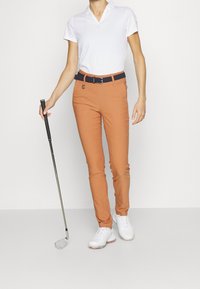Oranžové golfové kalhoty s úzkým střihem, doplněné námořnickým pleteným opaskem. Model má na sobě bílé polo tričko a bílé golfové boty, drží golfovou hůl.