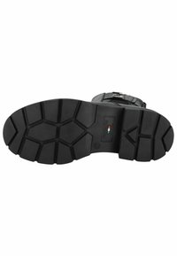 Semelle de botte en caoutchouc noir avec un motif de crampons profond. Présente des formes hexagonales et une étiquette de logo. Surface texturée pour une meilleure adhérence et durabilité.