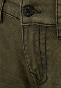 Olijfgroene denim jeans met een voorzak, donkere stiksels en een zwarte metalen knop met gegraveerde branding. Huidige structuur van de stof zichtbaar.