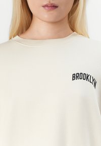 Sweatshirt crème avec un col côtelé et le texte "BROOKLYN" brodé en noir sur le côté gauche. Tissu doux avec une texture lisse.
